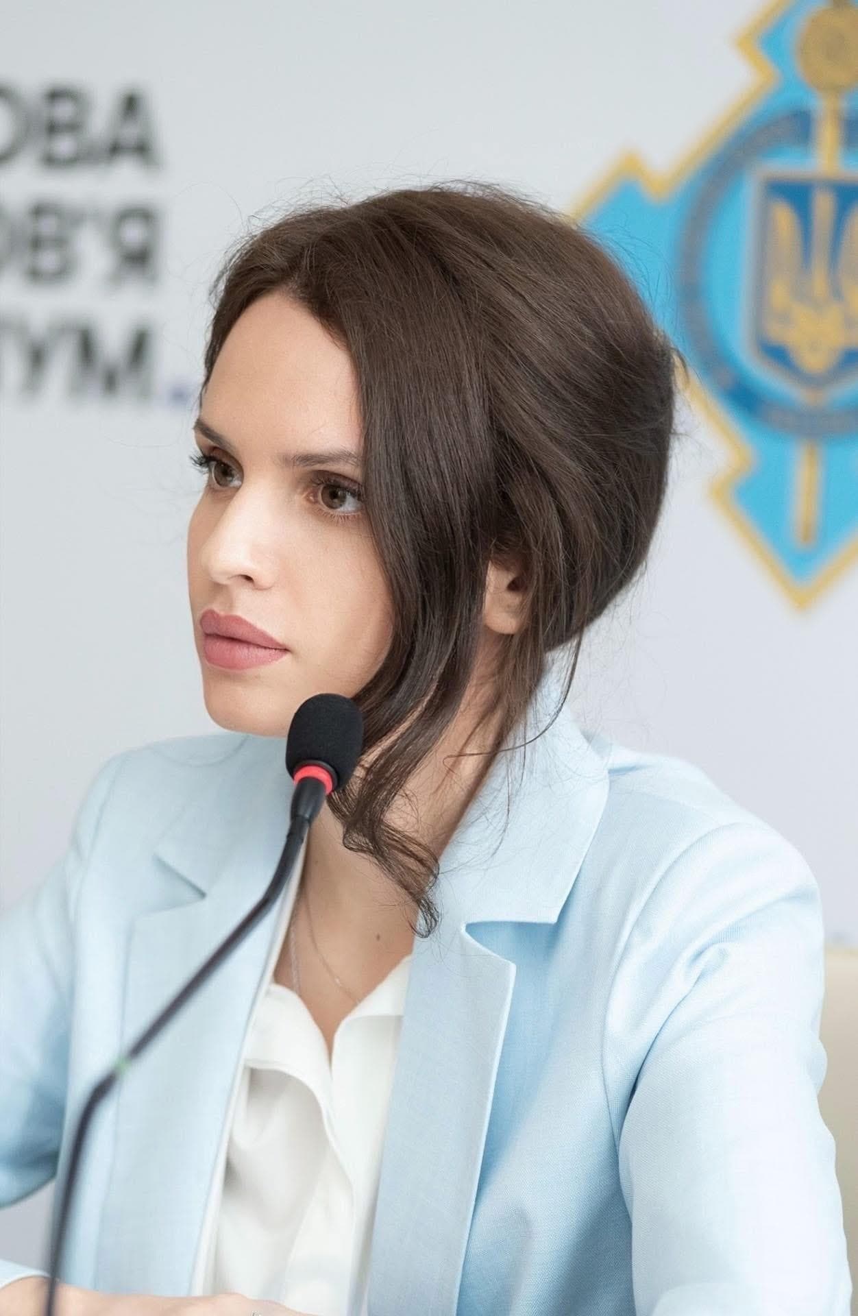 Anna Bon at a microphone on a panel, in a light-blue blazer, in front of a Ukrainian trident.