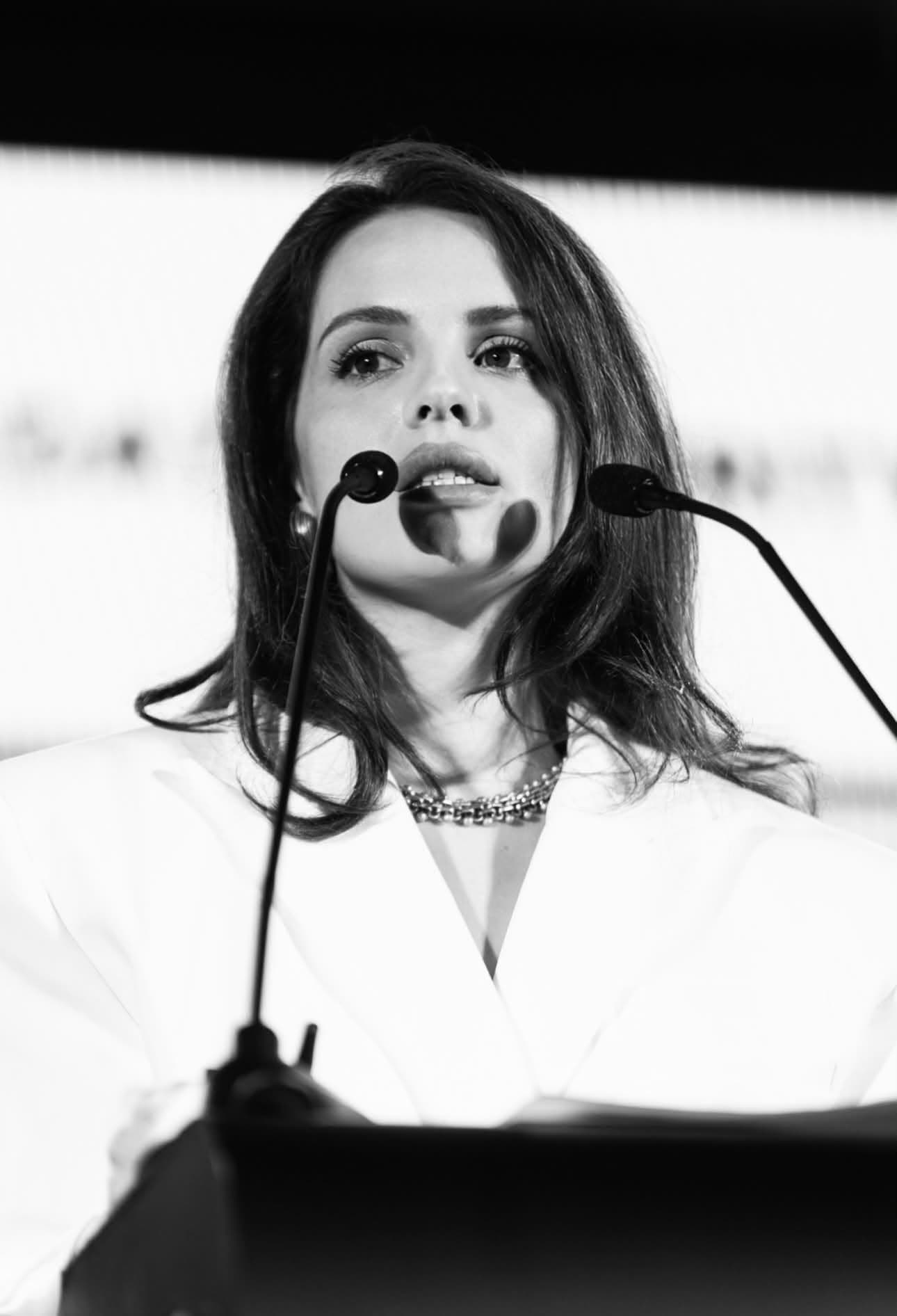 Anna Bon speaking at a podium behind two microphones, in black and white.